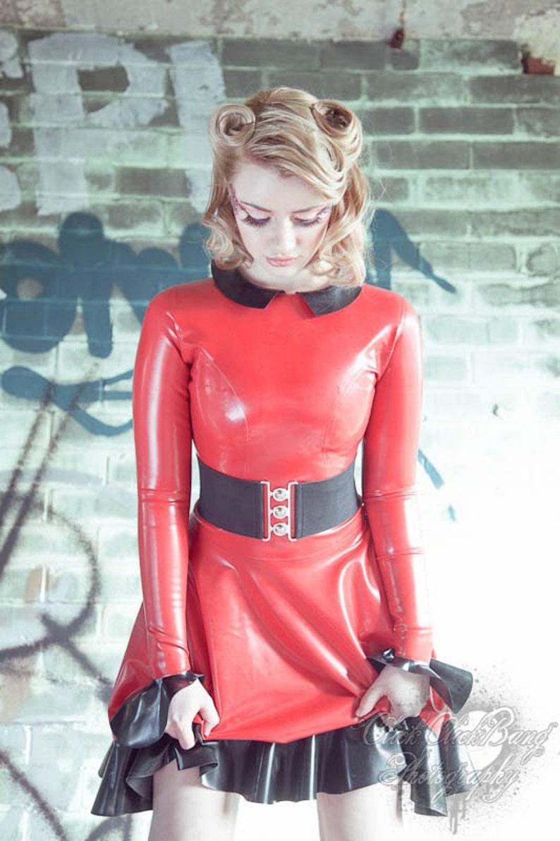 A pin up model wears a Fetasia Latex long sleeved swing dress. The dress is round necked with a black collar. The black flounced hemline is matched on the cuffs. The sleeves are and full length. The dress has a zip at the back.