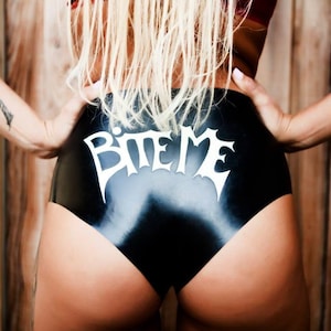 A blonde haired model wearing a black pair of shiny knickers. The pants feature a white "Bite Me" applique.