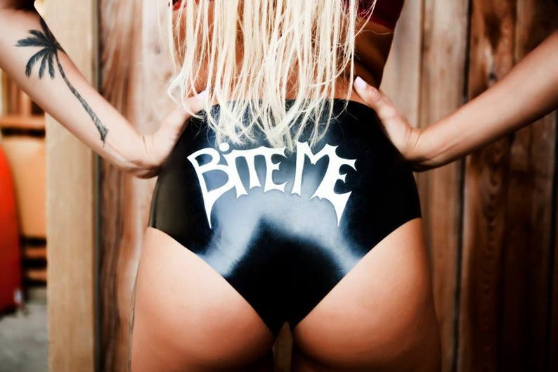 A blonde haired model wearing a black pair of shiny knickers. The pants feature a white "Bite Me" applique.