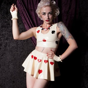 May include: A cream-colored latex crop top and pleated mini skirt set. The top and skirt are adorned with red heart and black spade suit symbols. Matching fingerless gloves with ruffled edges complete the outfit. A necklace with playing card suit symbols is also visible.