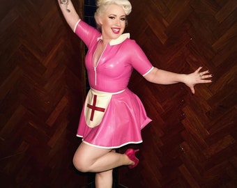 May include: A woman wearing a pink latex nurse costume with a white apron and a red cross. She is standing in a bathroom with wood paneling and black tile.