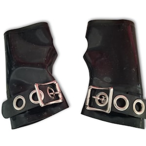 May include: Pair of black wrist cuffs. Each cuff features a buckle closure and a strap with three silver-colored eyelets. The cuffs are made of a shiny, possibly rubber or latex material.