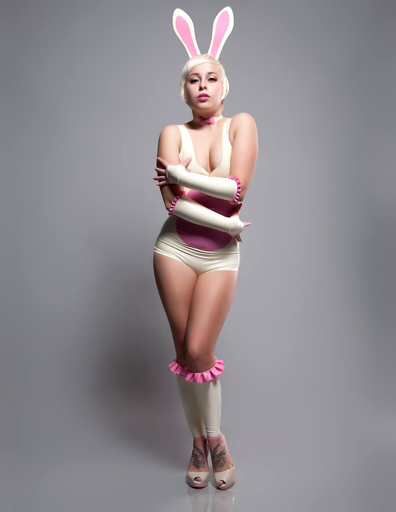 A woman wearing a white latex bunny costume with pink accents. The costume includes a bodysuit, gloves, and leg warmers. The woman is also wearing a pink bow tie and bunny ears.