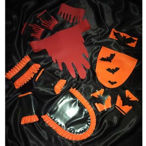 Può includere: Una collezione di accessori a tema Halloween in nero, arancione e rosso. Include un ritaglio rosso a forma di mano, ritagli arancioni a forma di pipistrello e pezzi rifiniti in nero e arancione. Gli articoli sono disposti su una superficie nera strutturata.
