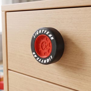 Pode incluir: Um puxador de gaveta circular em forma de pneu de corrida preto com uma jante vermelha no centro. A parede lateral do pneu preto apresenta o texto branco LIGHTYEAR impresso ao longo da borda. O puxador está fixado numa gaveta de madeira clara.