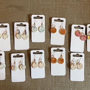 May include: A collection of handmade ceramic earrings displayed on white cards. The earrings feature various designs, including floral patterns and textured surfaces, in colors like white, orange, and pink. The earrings are suspended from silver-colored hooks.