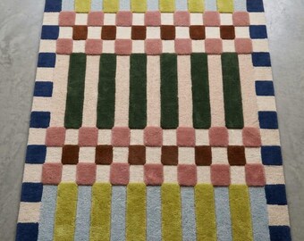 May include: A rectangular rug with a geometric pattern. The rug features alternating vertical stripes of green, blue, and beige, interspersed with rows of pink and brown squares. The border is a checkerboard of blue and beige squares.