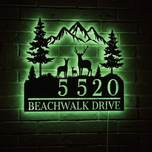Peut inclure: Panneau d'adresse en métal noir avec une silhouette de famille de cerfs, des arbres et des montagnes. Le panneau affiche les chiffres "5520" et le nom de la rue "BEACHWALK DRIVE". Il est éclairé d'une lueur verte sur un mur de briques.