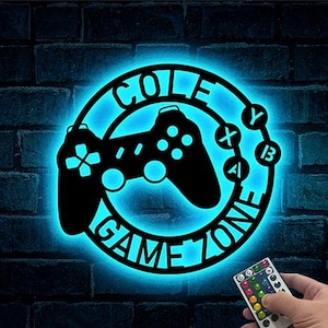 May include: A black metal "COLE GAME ZONE" sign with a glowing blue outline. The sign features a game controller design and the letters "Y", "X", "A", and "B". A hand holds a remote control with color options.