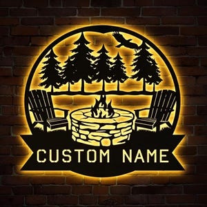 May include: A black metal sign with a backlit design featuring a campfire, Adirondack chairs, and pine trees. The sign includes the text "CUSTOM NAME" in a banner below the circular design, perfect for personalized decor.