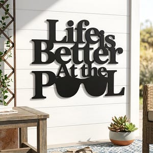 Könnte beinhalten: Schwarzes Metall-Wandbild mit dem Schriftzug "Life is Better At the Pool". Das Wort "Pool" wird durch eine Sonnenbrille ersetzt. Das Schild ist an einer weißen Wand mit horizontalen Brettern befestigt.