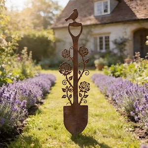 Könnte beinhalten: Eine dekorative, rostbraune Metall-Gartenschaufel mit einem Vogel obenauf. Die Schaufel hat ausgeschnittene Rosendesigns entlang des Stiels. Die Schaufel steht in einer Rasenfläche mit Lavendelbüschen auf beiden Seiten.