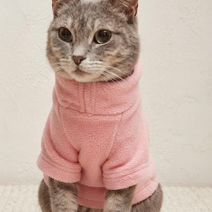 以下が含まれることがあります： ハイネックの柔らかいライトピンクのフリースセーターを着たグレーのトラ猫。セーターは半袖で、質感のある生地で仕上げられています。画像右上の隅には、MeowModelという文字が白でプリントされています。