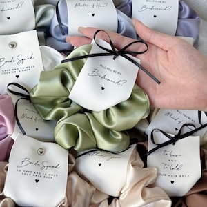 Könnte beinhalten: Eine Sammlung von Satin-Haargummis in verschiedenen Farben, die jeweils an einer weißen Karte mit einer schwarzen Schleife befestigt sind. Die Karten enthalten gedruckten Text wie Will You Be My Bridesmaid, Matron of Honor und Bride Squad mit Herzsymbolen.