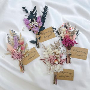 May include: Four small, dried flower bouquets with attached tags. The bouquets feature various colors, including pink, purple, and white, and are tied with twine. Each tag has a handwritten message: "Will you be my bridesmaid?", "Will you be my flower girl?", or "Will you be my matron of honour?"