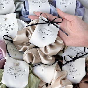 Könnte beinhalten: Eine Sammlung von Satin-Haargummis in verschiedenen Farben, jeweils an einer weißen Karte mit einer schwarzen Schleife befestigt. Die Karten enthalten gedruckten Text wie Will You Be My Bridesmaid, Maid of Honor und Bride Squad sowie kleine Herzsymbole.