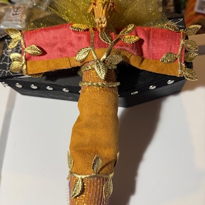 May include: A decorative, handcrafted item featuring a tan suede-like base, adorned with gold-colored leaf accents and a red and gold fabric top. The top is embellished with gold glitter and a detailed, animal-shaped centerpiece.