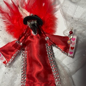 Può includere: Una figura di uccello in tessuto rosso fatta a mano con un cappello nero e ali di piume rosse. L'uccello è adornato con strass argentati e un gioiello a forma di cuore. La figura ha una bacchetta di perline con accenti a forma di cuore.