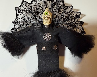 Op de afbeelding: Handgemaakte pop met een schedelkop en een enkel groen oog. De pop is gekleed in een zwarte outfit met een harige rand en een decoratieve spinnenwebachtige kraag. Op de borst zit een zilveren embleem.