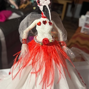 Könnte beinhalten: Eine handgefertigte Puppe mit einer vogelartigen Maske, verziert mit roten Herzakzenten. Das weiße Oberteil der Puppe hat rote Herzverzierungen, eine rote Rose und einen roten Federrock. Die Arme der Puppe sind mit weißem Netzstoff und roten Manschetten bedeckt.