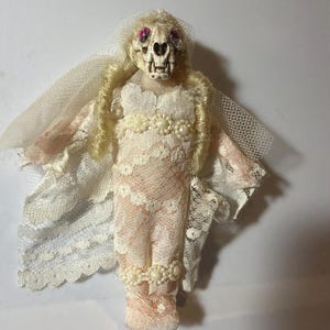 May include: A handcrafted doll featuring a skull head and pink eyes, dressed in a cream lace gown adorned with pearls. The doll has blonde hair and a veil, embodying a distinctive gothic bride theme.