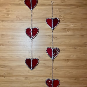 Puede incluir: Dos conjuntos de adornos colgantes con forma de corazón. Cada conjunto presenta tres corazones de vidrieras rojas conectados por una cadena plateada. Los corazones tienen un borde plateado y están suspendidos sobre un fondo de madera.