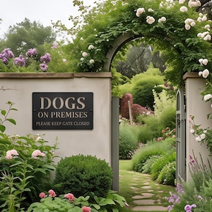 May include: A rectangular black sign with white text reading DOGS ON PREMISES PLEASE KEEP GATE CLOSED, mounted on a light-colored stone wall next to a garden gate surrounded by lush green foliage, white climbing roses, and purple flowers.