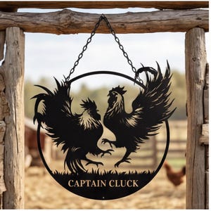 Könnte beinhalten: Schwarzes Metallschild mit zwei sich gegenüberstehenden Hähnen in einem kreisförmigen Rahmen. Das Schild wird von einer Kette gehalten und trägt den Text "CAPTAIN CLUCK" am unteren Rand. Der Hintergrund zeigt einen Holzzaun und eine verschwommene Außenszene.