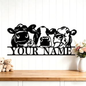 May include: Black metal wall art featuring three cow faces with the text "YOUR NAME" below. The cows have detailed features and are set against a white background. A flower is between two of the cows.