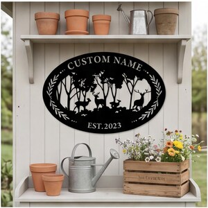 May include: An oval black metal wall sign featuring a laser-cut forest scene with deer silhouettes. The text CUSTOM NAME is arched at the top, and EST. 2023 is centered at the bottom. The design is framed by decorative leaf patterns on both sides.