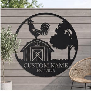 Op de afbeelding: Zwarte metalen cirkelvormige wandkunst met een haan op een schuur, een boom en de tekst "CUSTOM NAME EST 2025". Ontworpen voor buitengebruik, gemonteerd op een houten lattenwand.