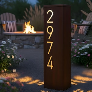 Peut inclure: Un panneau d'adresse rectangulaire en métal brun avec les chiffres 2974 illuminés d'une lumière dorée. Le panneau est installé dans un jardin avec un foyer, des chaises et des guirlandes lumineuses, créant une ambiance extérieure chaleureuse.
