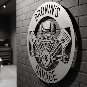 May include: A silver metal garage sign with the text "BROWN'S GARAGE" in a circular design. The sign features an engine design with pistons and a wrench, mounted on a dark gray brick wall. A classic car is visible in the background.