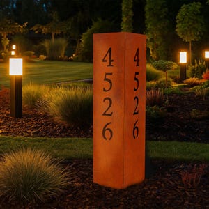 Könnte beinhalten: Ein hoher, rechteckiger Adressschild aus verwittertem, rostfarbenem Metall. Die Zahlen "4526" sind dunkel ausgeschnitten, sodass Licht hindurchscheinen kann. Das Schild steht in einem Garten mit sanfter Beleuchtung.