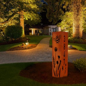 May include: An orange metal address sign featuring a bee and floral designs. The numbers "3847" are cut out vertically. The sign is illuminated at night, set in a garden with a pathway leading to a house.