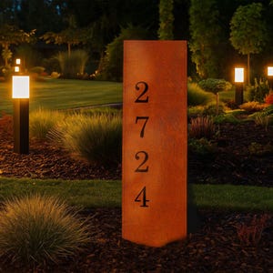 Könnte beinhalten: Ein rechteckiges, verwittertes Adressschild aus Metall mit den schwarzen Zahlen 2724. Das Schild hat einen rostigen Orangefarbton und befindet sich in einem Garten mit grünem Gras, Büschen und kleinen Lichtern.