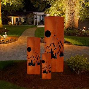 Può includere: Tre sculture in metallo da esterno con finitura arancione ruggine. Ogni scultura rettangolare presenta un design di montagna e alberi neri con un ritaglio circolare. Le sculture variano in altezza e sono collocate su un letto di pacciame in un giardino.