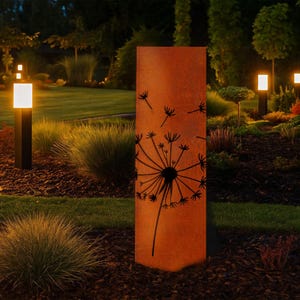 Op de afbeelding: Een hoog, rechthoekig tuin kunstwerk van roest-oranje metaal met een zwart paardenbloem ontwerp. Het kunstwerk staat in een tuin met groen gras, struiken en verlichte padverlichting.