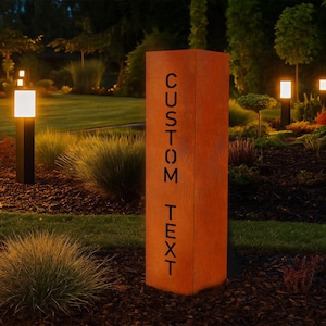Op de afbeelding: Een hoge, rechthoekige, roest-oranje metalen paal met de woorden "CUSTOM TEXT" in zwart, verticaal uitgelijnd. De paal staat in een tuin met verlichte padverlichting en weelderig groen.