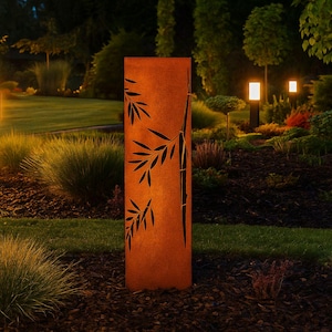 May include: A tall, rectangular, rusty-orange metal garden sculpture featuring a black bamboo leaf design. The sculpture stands in a garden with green grass, shrubs, and illuminated pathway lights. The overall scene is set at dusk.