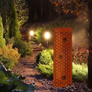 May include: A tall, rectangular, rusty-orange metal garden art piece featuring a honeycomb pattern with three bee silhouettes. The art piece is illuminated by garden lights, creating a warm ambiance in a garden setting.