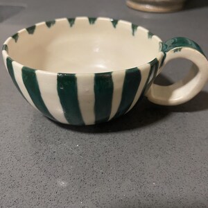 May include: A ceramic teacup with a white interior and exterior, featuring vertical dark green stripes. The handle is also dark green. The rim has a scalloped edge. The cup sits on a grey surface.