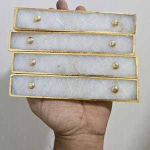 Puede incluir: Un conjunto de cuatro tiradores rectangulares apilados. Cada tirador presenta una cara de mármol blanco con un borde dorado y un pomo dorado. Se sostienen en una mano sobre un fondo blanco liso.