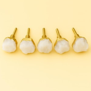 Puede incluir: Cinco tiradores decorativos para armarios con un vástago dorado y una piedra blanca de forma irregular. Los tiradores están dispuestos en fila sobre un fondo amarillo claro. Adecuados para muebles y decoración del hogar.