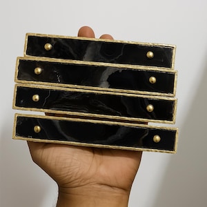May include: Four rectangular drawer pulls with a black marble-like design and gold trim. Each pull has a single gold-coloured knob. The pulls are held in a hand against a white background. Home decor.