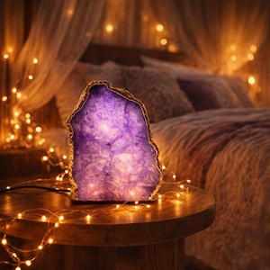 May include: A purple agate lamp with a gold-colored edge sits on a wooden table. The lamp is lit, and the light shines through the stone. A string of small, warm white lights surrounds the table, and a bed is in the background.