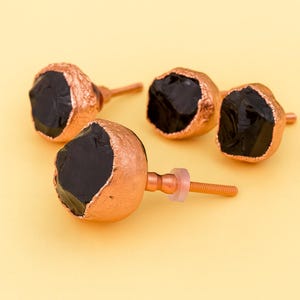 May include: Four cabinet knobs featuring a raw black stone centre and copper-coloured metal trim. The knobs are set against a soft yellow background, highlighting their unique, organic design. Each knob has a screw for easy installation.