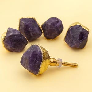 Puede incluir: Cinco tiradores de armario de amatista con bordes chapados en oro. Los tiradores tienen un aspecto natural y tosco, mostrando el color púrpura intenso de la amatista. Un tirador está completamente montado, mostrando el tornillo y los herrajes.