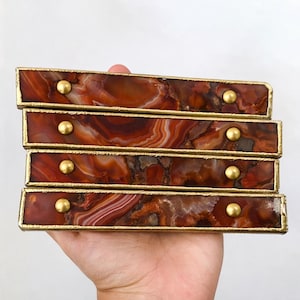 May include: Four rectangular decorative drawer pulls with a rich, reddish-brown agate pattern. Each pull has a gold-coloured metal frame and a single, round, gold-coloured knob. The pulls are stacked and held in a hand.