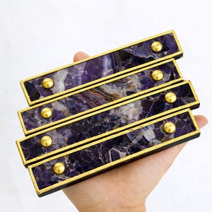 May include: Four rectangular cabinet pulls with a deep purple and white marbled design. Each pull has a gold-colored frame and gold-colored round accents. The pulls are held in a hand against a white background.